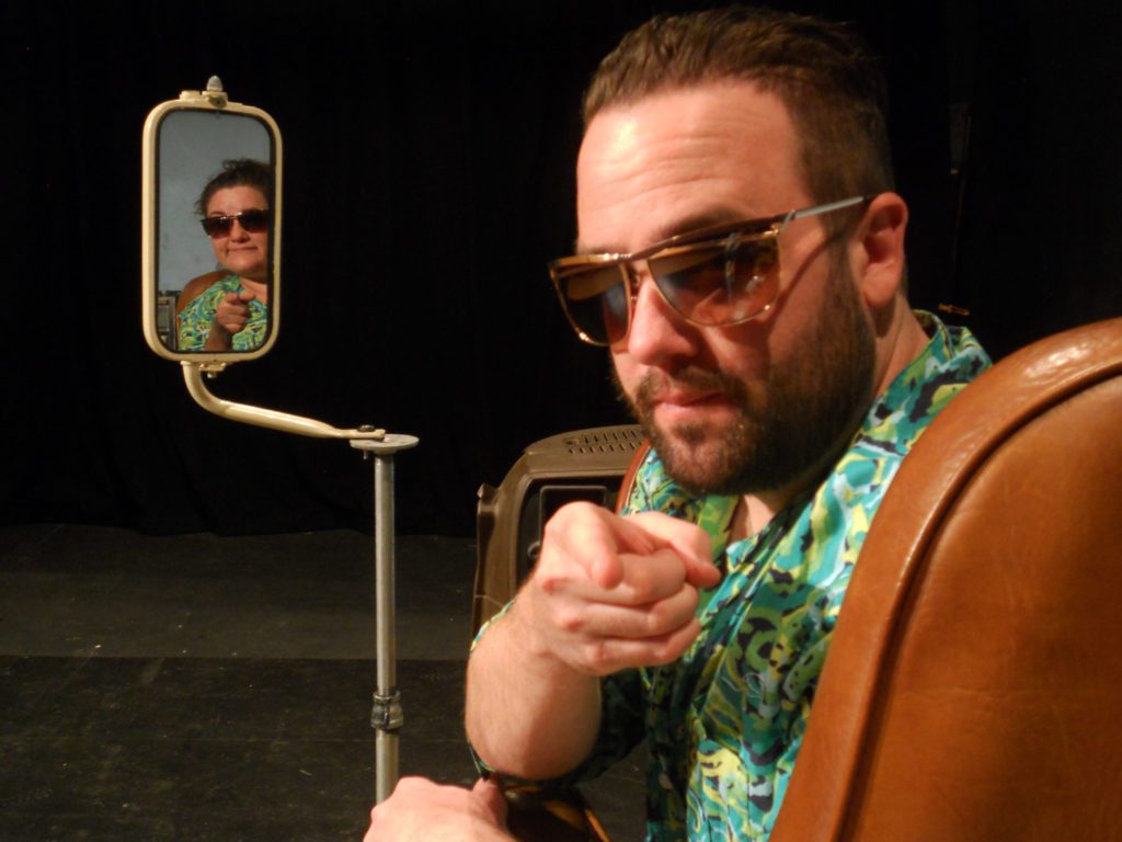 A man with sunglasses and a brightly colored shirt sits in a brown driver's seat, twisting to stare seriously at the camera. Next to him, in a driver's side mirror, is a woman in the same sunglasses. Both are pointing