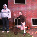 A lady wearing jeans and a sweatshirt hoodie that says “Property of Lasagna” with a picture of Garfield on it. She looks upset. Next to her is hole that had been dug into the dirt and a man is down on one knee trying to explain something to her.