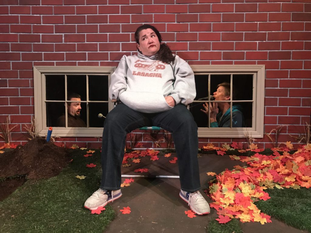 A woman is sitting in her backyard in an old folding chair. Behind her in the basement windows of the house, you can see a man in the window on the right and his best friend in the window on the left and they are in the middle of recording a pod cast.