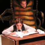 A man writing at a desk concentrates as a large beetle looms behind him.
