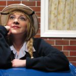 Close-up of a postal worker with long blond braids leaning on a blue mailbox while smiling. In the background is a brick wall with a curtained window.