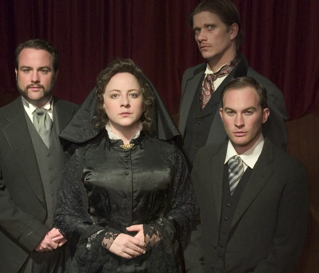 A woman and three men in 1800s funeral attire are looking forward in front of a red curtain.