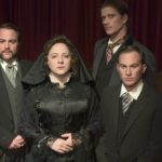 A woman and three men in 1800s funeral attire are looking forward in front of a red curtain.