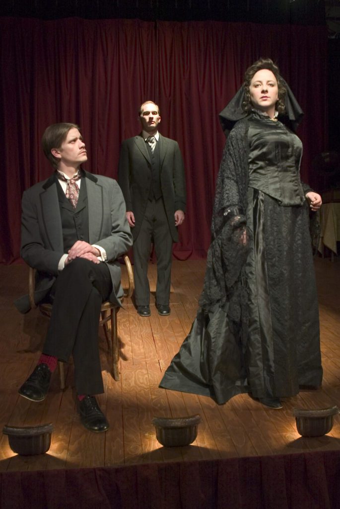 A man in a black 1800s suit is seated in a wooden chair facing forward with his head looking to his left at a woman in a black funeral dress. they are on a wood planked floor with foot lights and a red curtain behind in the distance. Between them stands another man in dramatic overhead light.