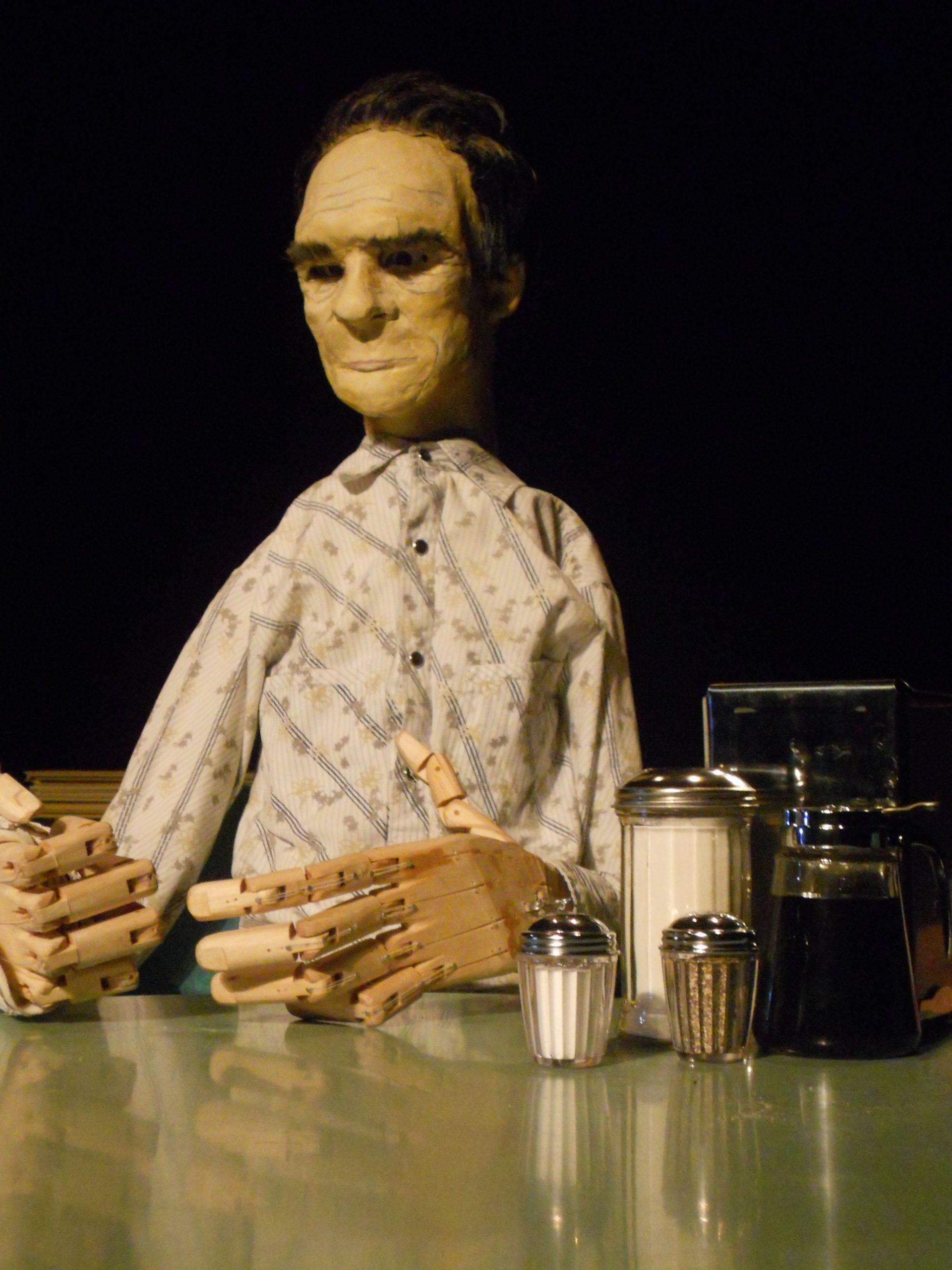 Close up of a life-size puppet of Tommy Lee Jones sitting at a diner table. His hands are wooden and are in the foreground. They are built to work the same way as human hands and you can see the mechanism is uses fishing line and rubber bands.