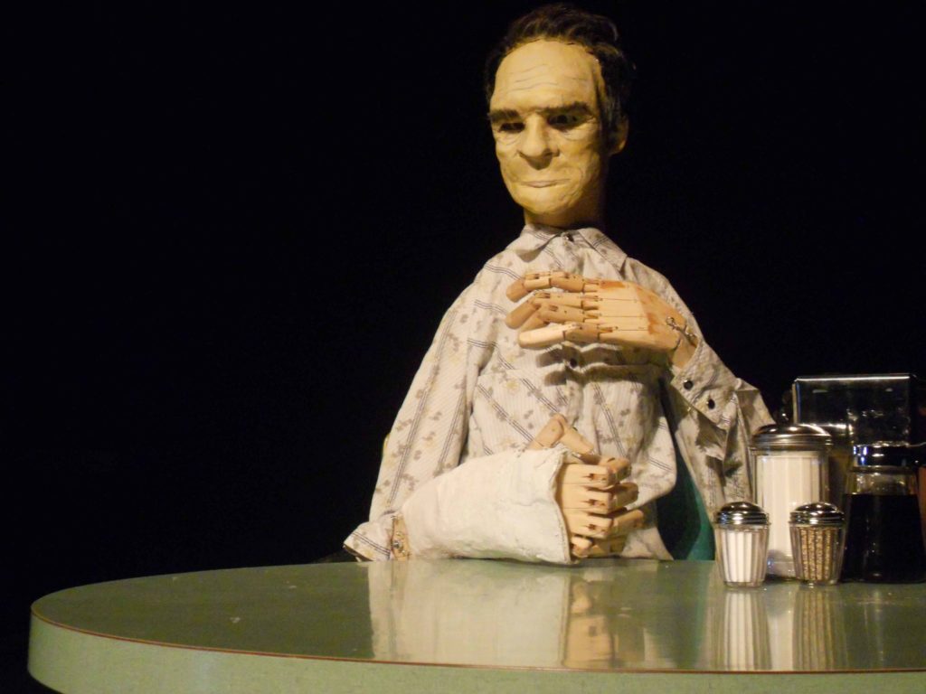 The life-size puppet of Tommy Lee Jones is sitting at a diner table. His left hand, made of wood, is held to his chest and his right arm is resting on the diner table.
