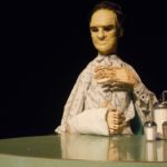 The life-size puppet of Tommy Lee Jones is sitting at a diner table. His left hand, made of wood, is held to his chest and his right arm is resting on the diner table.