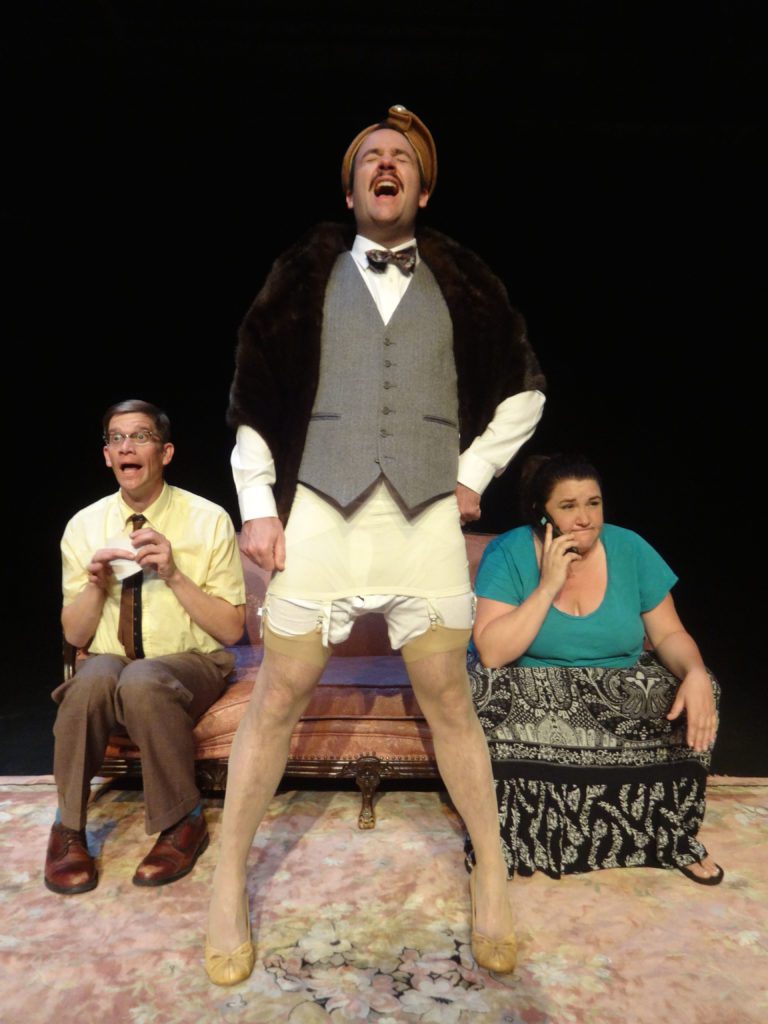 A man stands with his head back singing or yelling, perhaps. He wears a suit on the top and stockings and high heels on the bottom. Behind him, two people are seated on a pink couch. One is a man in a yellow shirt talking excitedly. The other is a woman in a blue t-shirt looking on a cell phone looking annoyed.