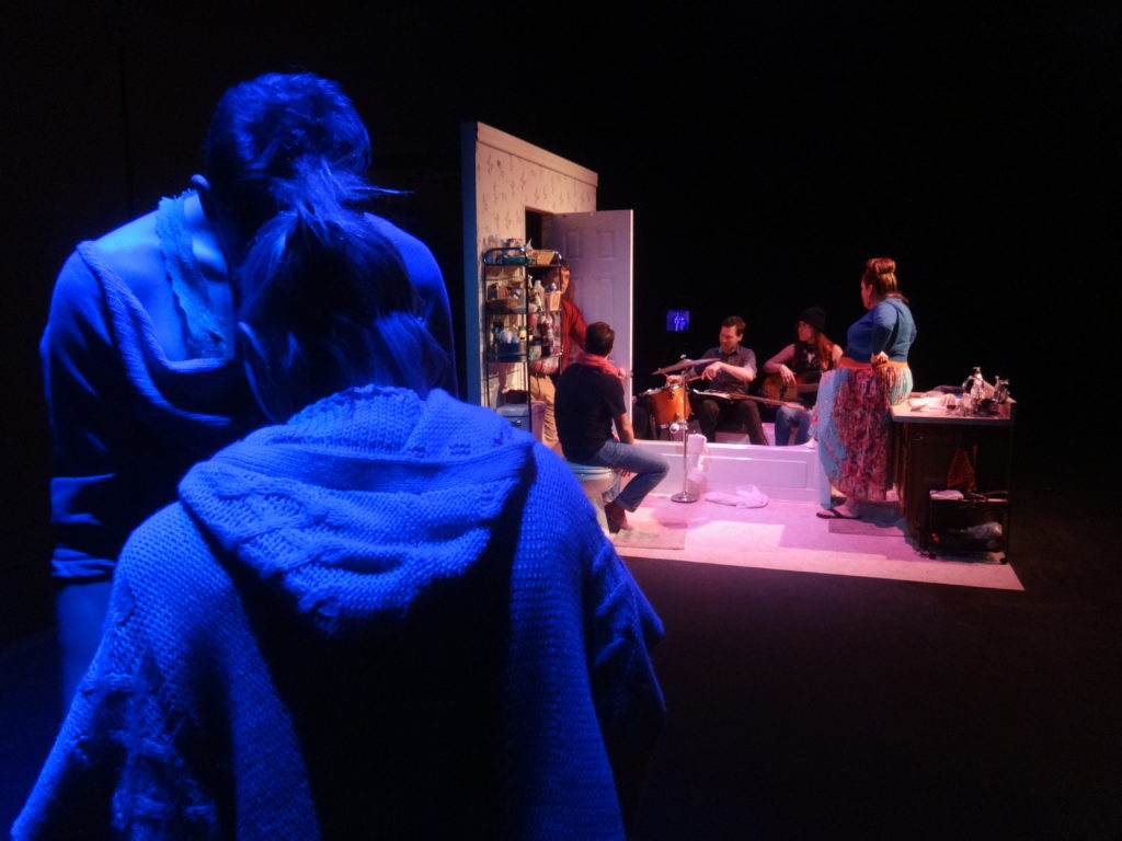 In the foreground two people dance in a dim blue light. Behind them a group of people stand in a small bathroom. A band plays in the bathtub.