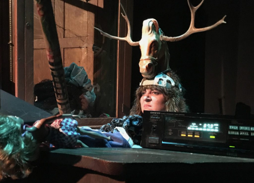 Peaking up from behind a bunch of clutter is the top of someone's head. They are glaring at something in the distance and have a large headpiece that includes a plastic horse head with antlers.