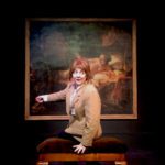 A female art museum security guard sits on an upholstered bench in front of a life size nude painting of Danae (the mother of Perseus) by Rembrandt. She’s speaking out and gesturing back to the painting.