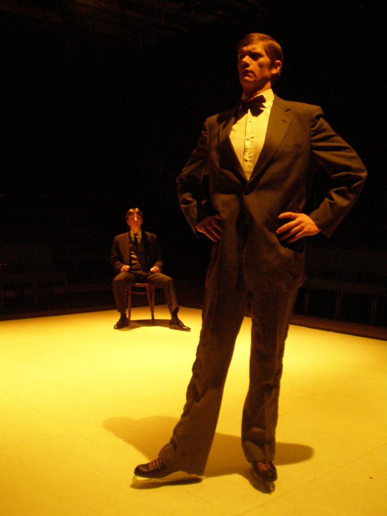 A man in stark lighting poses in the foreground while wearing ice skates. In the background, another man sits in a chair staring at him.
