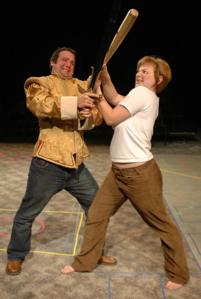 On a carpeted floor, two people are in a battle. One has a sword, the other has a baseball bat.