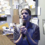 A man sits at a desk singing earnestly into a microphone. The walls behind him are covered in papers, letters, and postcards.
