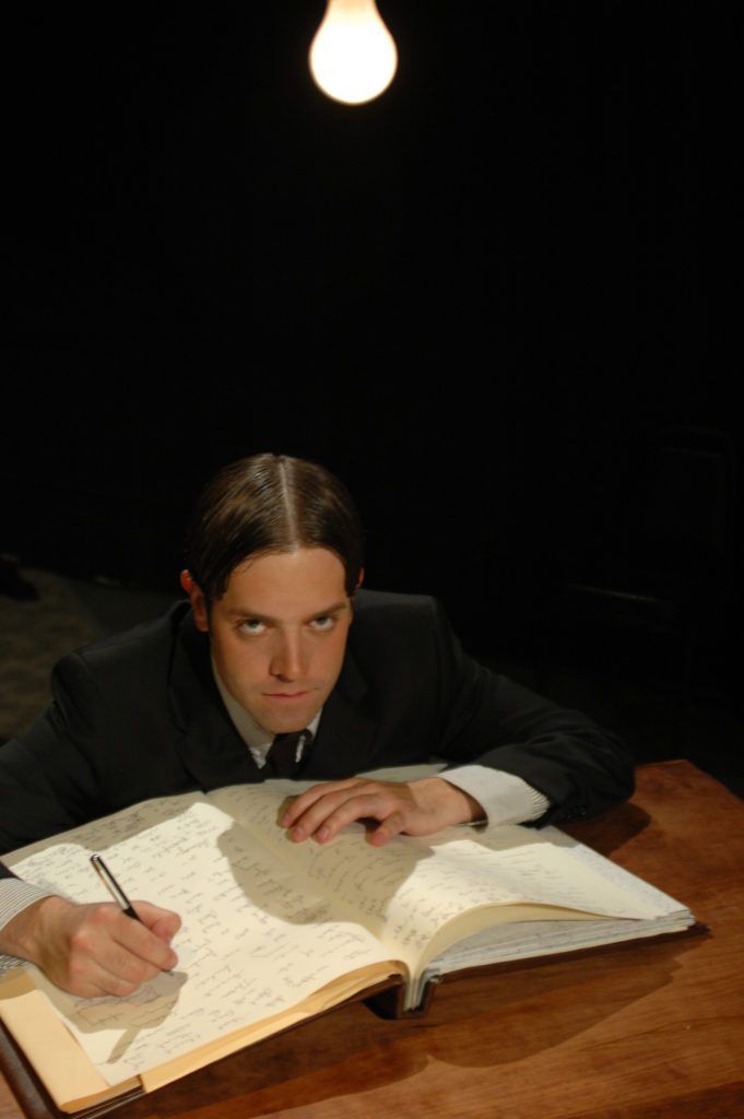 A serious man looks up from a desk where he is writing. A single bare lightbulb hangs above.
