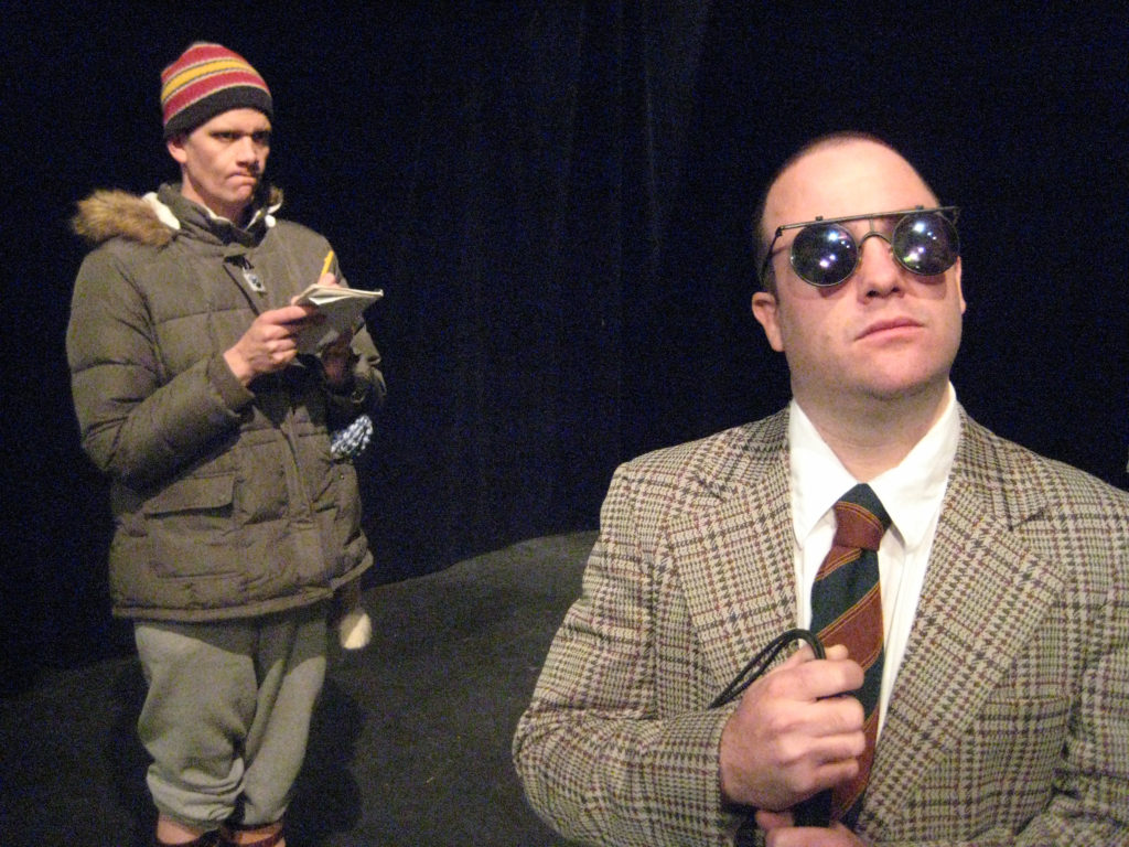 A man in dark glasses and a patterned suit stands in the foreground. Behind him, a man in a winter coat and hat stares furiously at the him, while writing in a notebook.