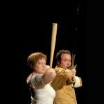 A woman, her body facing sideways but looking at the camera poses with elbows up holds a bat straight up near her face. A man right in front of the woman holds a sword in the same position