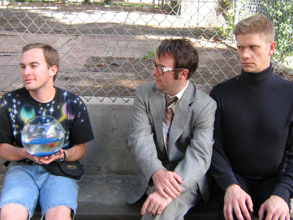 Three awkward men sit in front of a chainlink fence. On the left is a smiling man with a space t-shirt on he is holding a fish bowl with a goldfish. In the center, a man in a suit and large glasses purses his lips. On the right is an intense man with a buzz cut and black turtleneck.