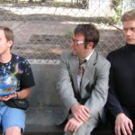 Three awkward men sit in front of a chainlink fence. On the left is a smiling man with a space t-shirt on he is holding a fish bowl with a goldfish. In the center, a man in a suit and large glasses purses his lips. On the right is an intense man with a buzz cut and black turtleneck.