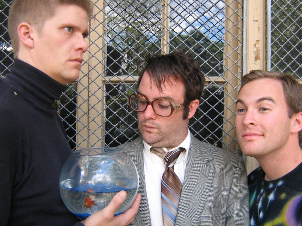 Three awkward men in front of a window grate. On the left is an intense man with a buzz cut and black turtleneck, holding a fish bowl with a goldfish in it. In the center, a man in a suit and large glasses purses his lips. On the right is a smiling man with a space t-shirt on