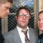 Close-up of three awkward men in front of a window grate. On the left is an intense man with a buzz cut and black turtleneck. In the center, a man in a suit and large glasses purses his lips. On the right is a smiling man with a space t-shirt on