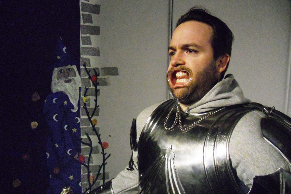 A man dressed in a suite of armor stands in front of a painting of a wizard. The man is wearing the lip expanders you might wear at the dentist’s office during a procedure.