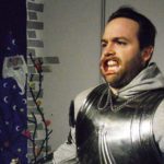 A man dressed in a suite of armor stands in front of a painting of a wizard. The man is wearing the lip expanders you might wear at the dentist’s office during a procedure.