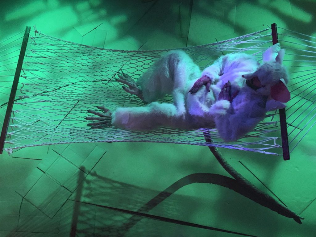 Looking down from above, we see a giant white Rat lying in a rope hammock rubbing his big old belly in green light.