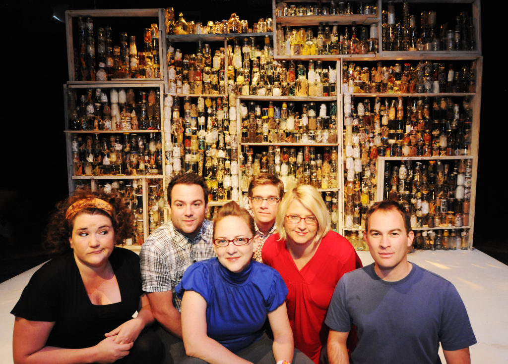 Six people are in the foreground. Most of them are smiling, but one is giving a grimacing side eye to the rest. In the background is a large wall full of hundreds of clear jars, which are all full of different items. It is the set of a Buntport play titled “Indiana, Indiana”.