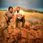 Two people are standing, knees bent in action. The woman is angrily talking, the man looks confused and surprised. They are standing a dry prairie scene, where a taxidermied badger and rattlesnake are facing off. The backdrop is a painted scene with distant mountains.