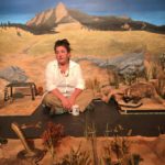 A woman in a white shirt sits cross-legged on the floor, looking despondent. She has a coffee cup in front of her. She is surrounded by an unfinished prairie scene, including a taxidermied badger facing off with a taxidermied rattlesnake. Behind all of them is a painted backdrop with a distant mountain.