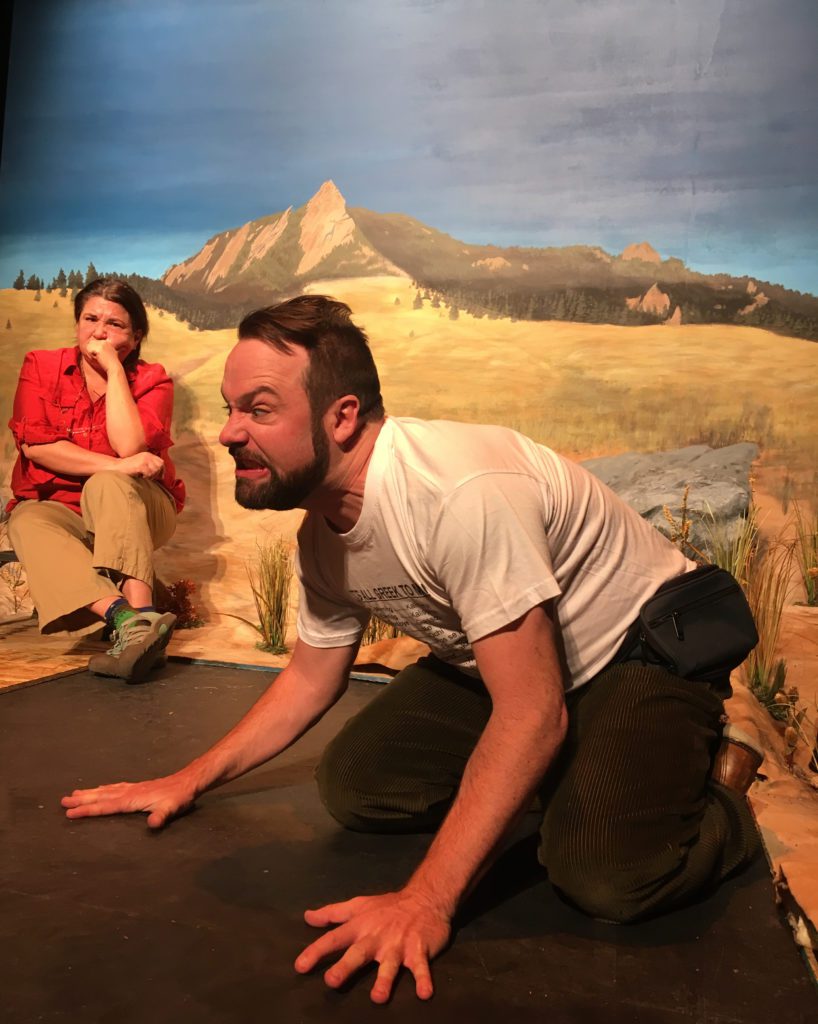 A bearded man is on all fours, his face scrunched up in anger. In the background a woman in a bright orange shirt sits on a small stool looking on, angry or bored or both. Behind them is a painted backdrop with a distant mountain.