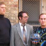 Three awkward men in front of a window grate. On the left is an intense man with a buzz cut and black turtleneck. In the center, a man in a suit and large glasses purses his lips. On the right is a man with a space t-shirt on he is holding a fish bowl with a goldfish in it