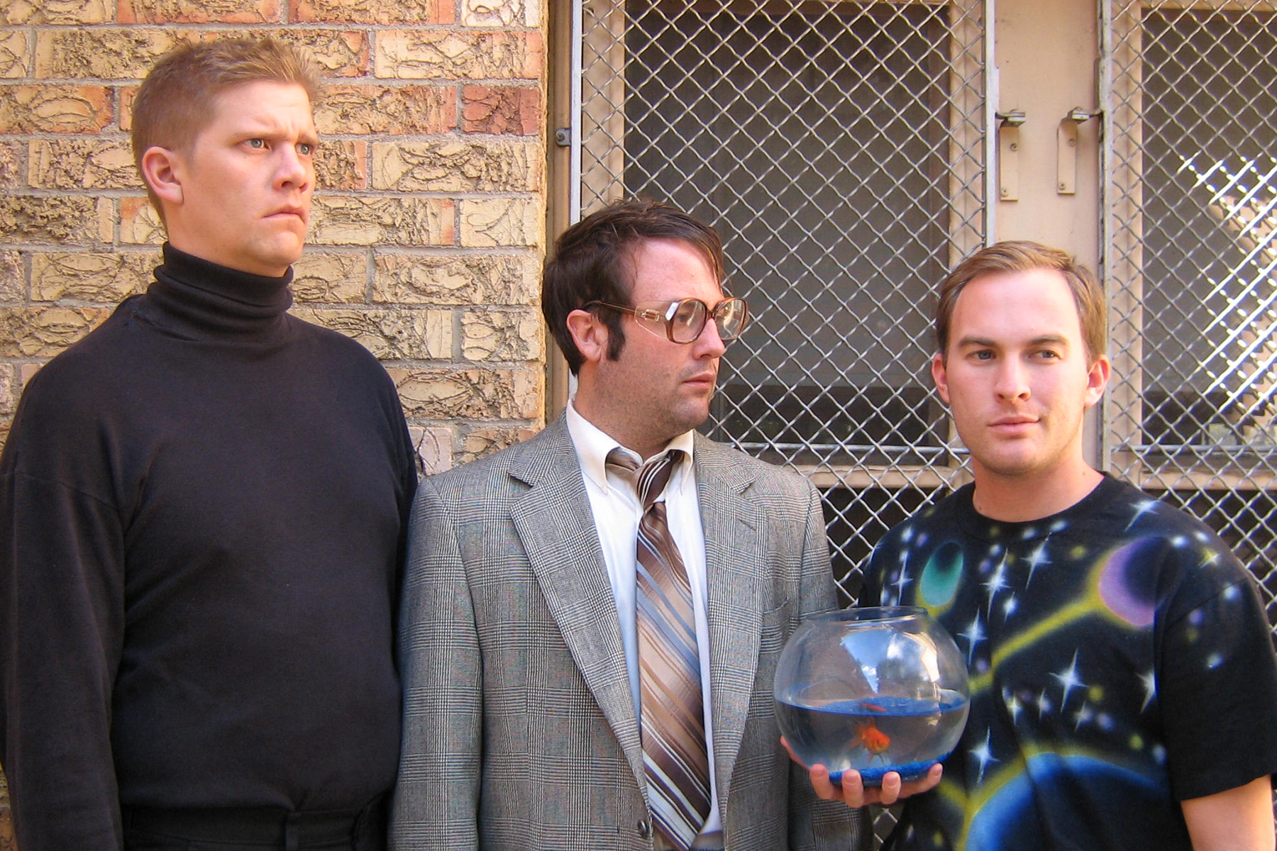 Three awkward men in front of a window grate. On the left is an intense man with a buzz cut and black turtleneck. In the center, a man in a suit and large glasses purses his lips. On the right is a man with a space t-shirt on he is holding a fish bowl with a goldfish in it