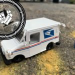 A tiny mail truck and a tinier blue mailbox are in a parking lot. Close-up of yellow paint lines are in the foreground. Stamped in the upper left corner is a black-and-white seal that says “Buntport Bored Post Society Society”. In the center of the seal is a mailbox with some letters.