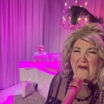 In the foreground, a sobbing blond woman is at a gold microphone. In the background, a man in a white suit sits at a white baby grand piano. The stage is washed with bright pink light.