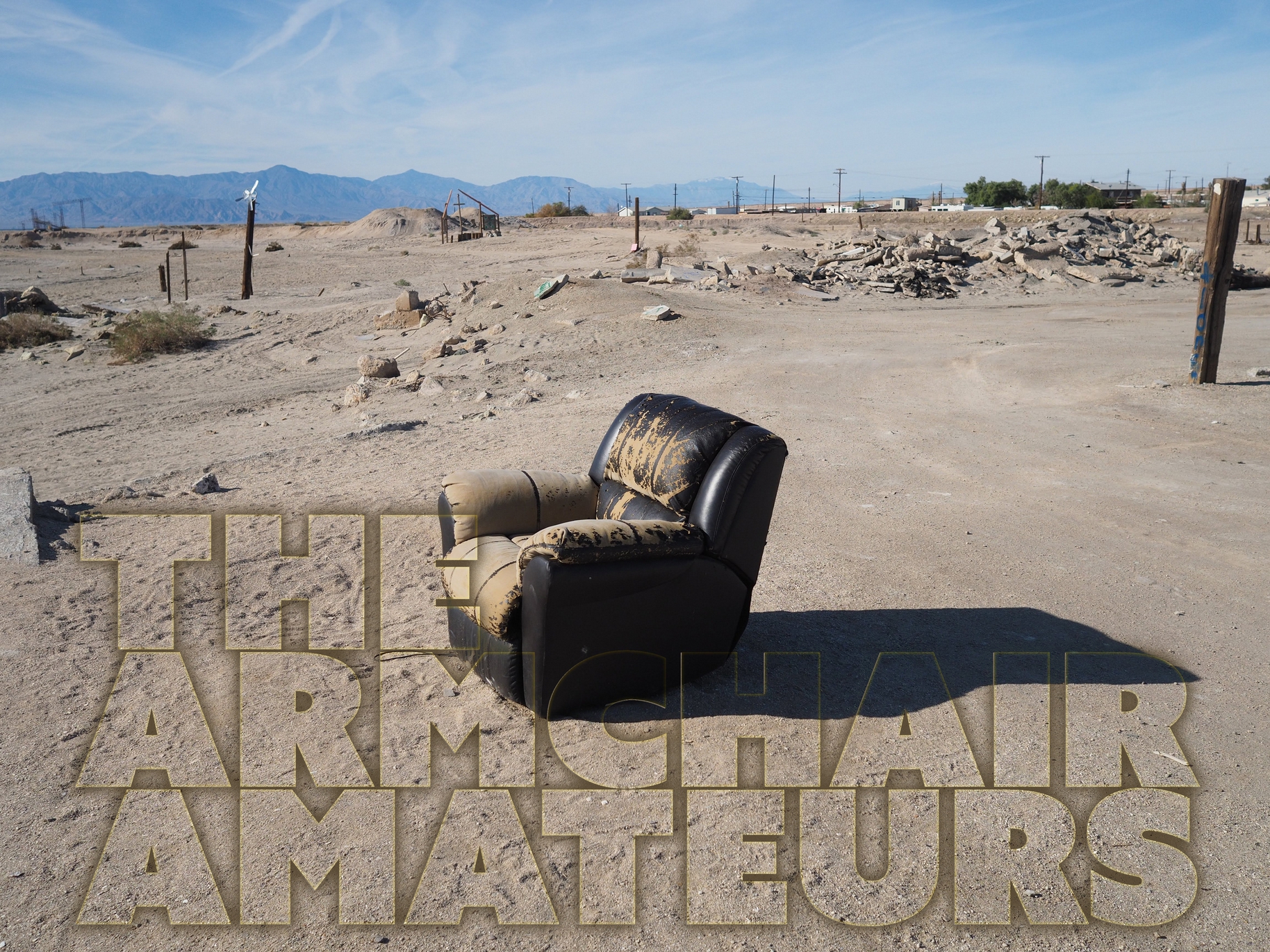 A photo of a worn down leather armchair in the middle of a desert, with telephone poles in the distance and the words "The Armchair Amateurs" in the lower left corner.