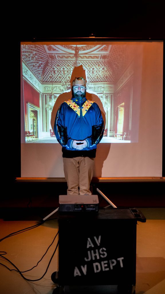 A man wearing what looks like a homemade regal looking jacket and a brown paper crown stands in front of a screen that has a royal church interior projected on it.