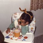 A baby, with antlers and black deer nose, sits in a highchair with a cup of tea.