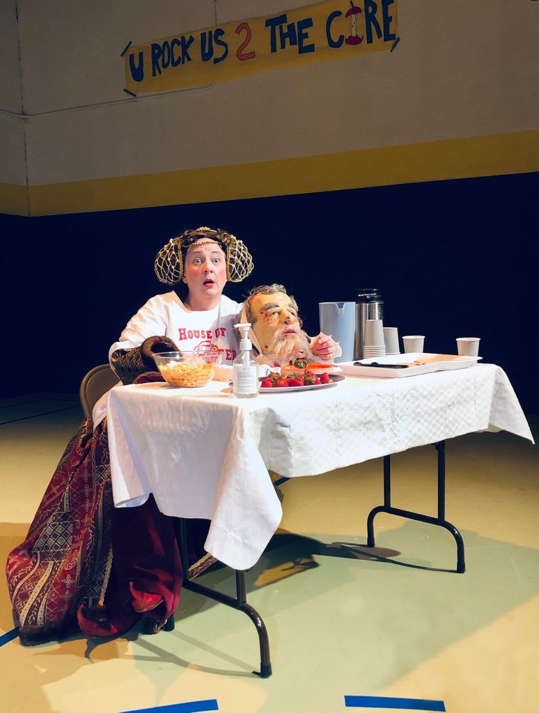A woman dressed in 15th Century clothing with a giant t-shirt over it sits at a table with snacks and a severed head on it.