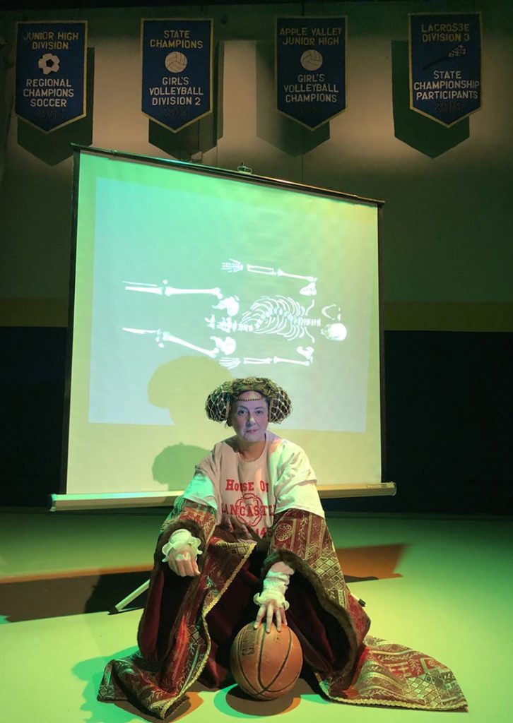 A woman dressed in 15th Century clothing with a giant t-shirt over it kneels with a basketball in front of a screen showing a picture of the crooked skeleton of Richard III.