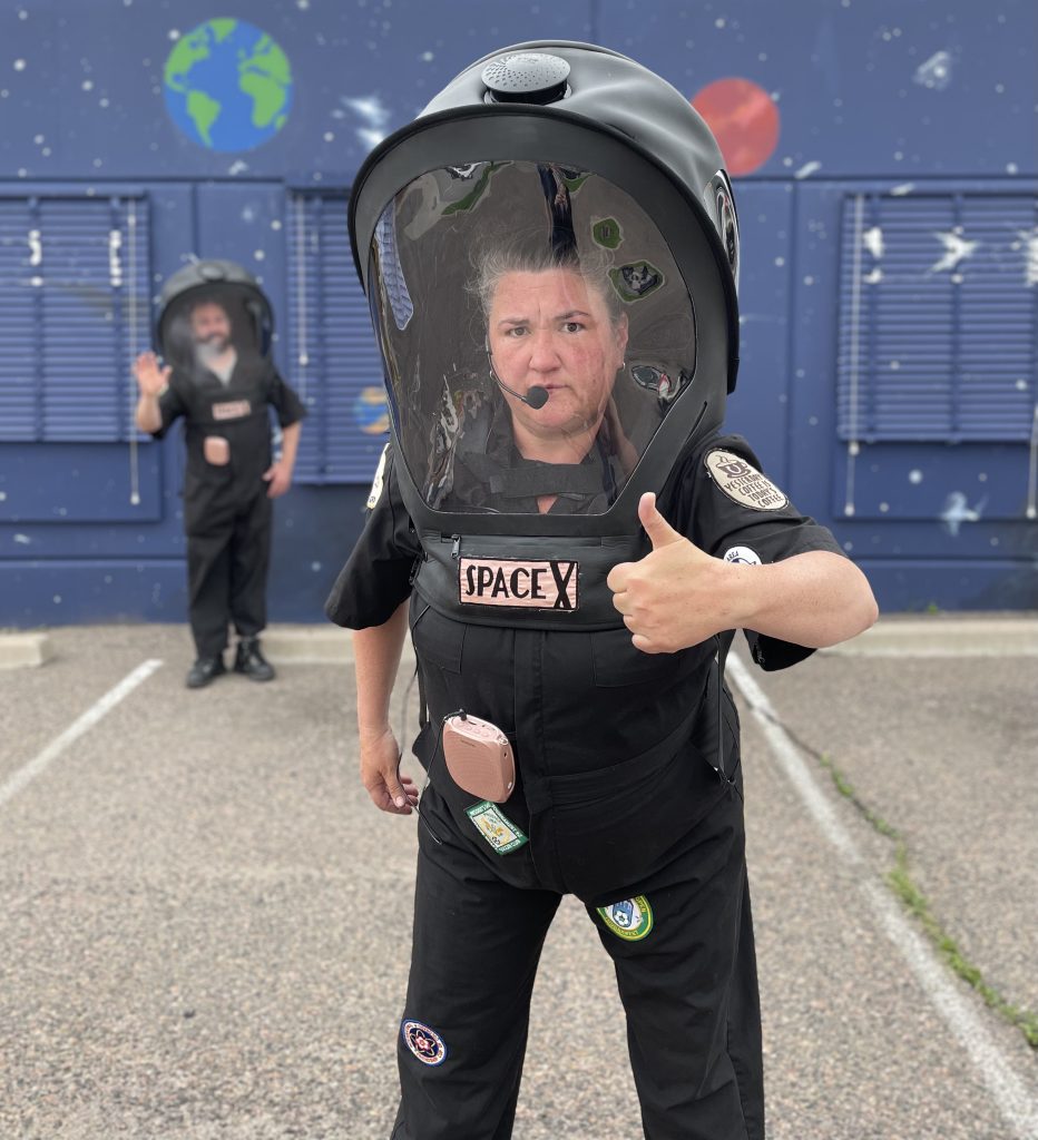 A close up of a space person wearing a huge black space helmet who is bending over and giving a thumbs up to the person viewing the picture. In the blurry distance we can see another space person waving to whoever is looking.