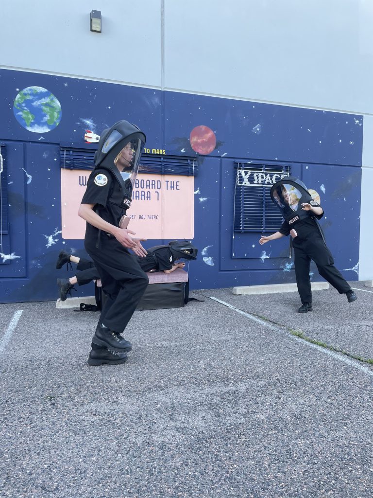 Three space people wearing huge black space helmets are in a parking lot in front of a wall that has been painted to look like outer space. The two people in the foreground look as if they are trying to look like they are floating and a third space person is laying on their belly on a bench as if they are floating.