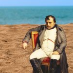 Napoleon Sits slumped over in a red chair, one a beach with water in the background. he looks unhappy.