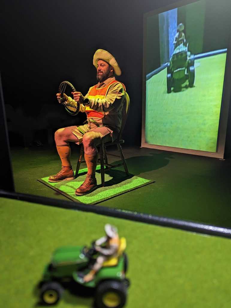 In the center is a man with a beard wearing a safety vest and a floppy hat. He is seated and holding a steering wheel. In the foreground is a miniature version of the man on a tractor. Behind The man is a projection of himself as the miniature.