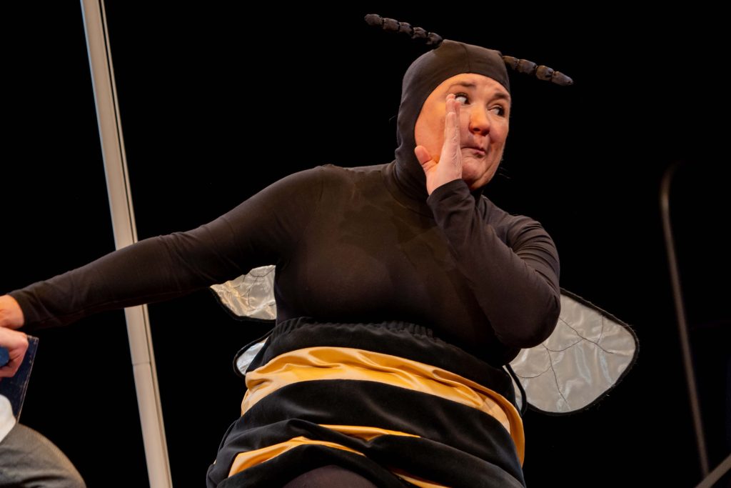A person dresses as a bee whispers with hand to face.