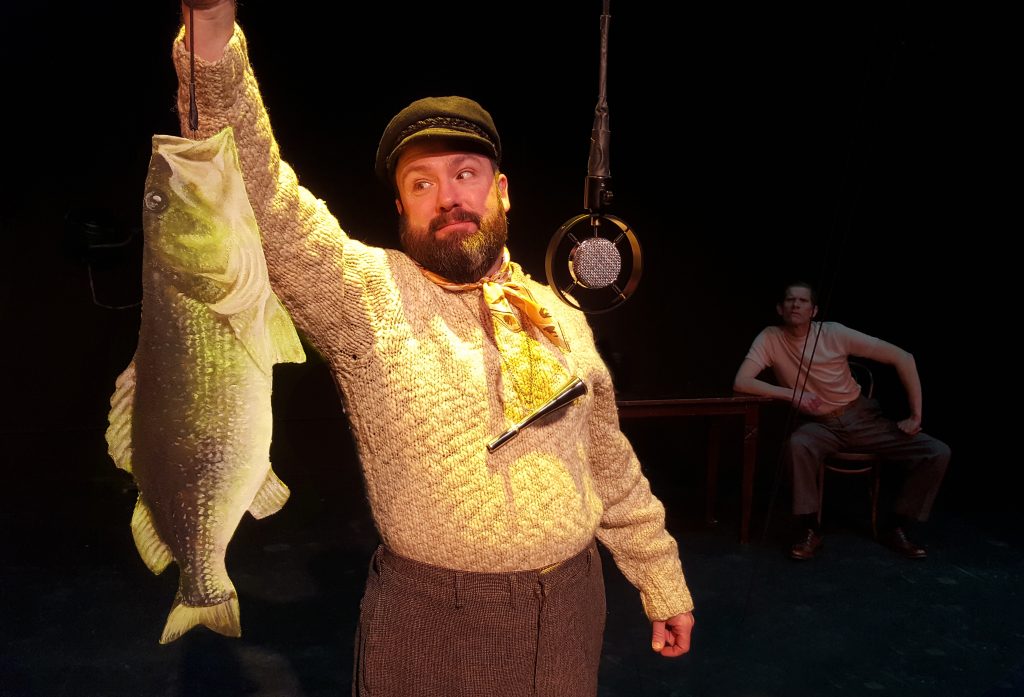 A man in a sweater and a cap holds up a 2D fish on a line, as though he has caught it. In front of him is a vintage microphone and, in the distance, another man sits, dimly lit and staring at the first man.