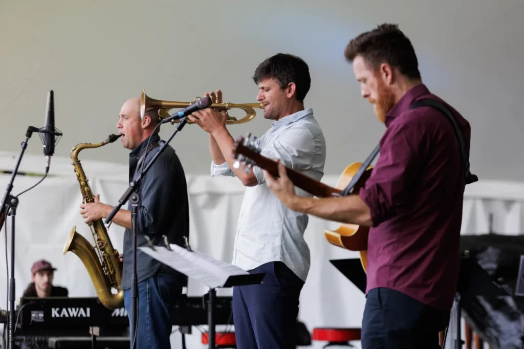 3 men stand at microphones, palying instruments.