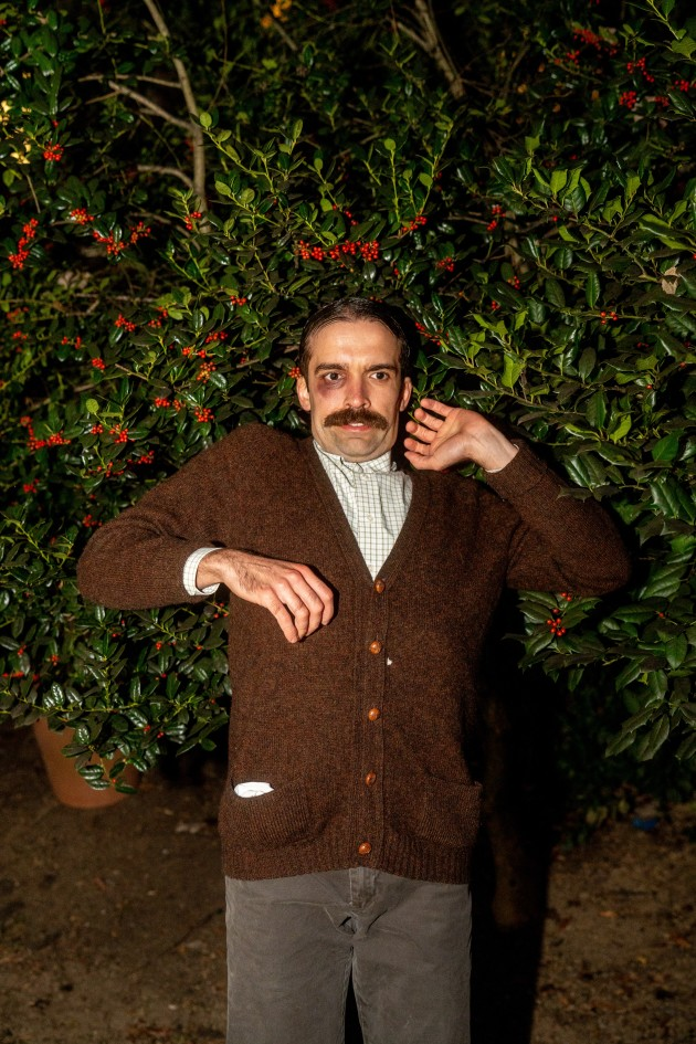 A man with a black eye and mustache is laying on flowers and greenery with a