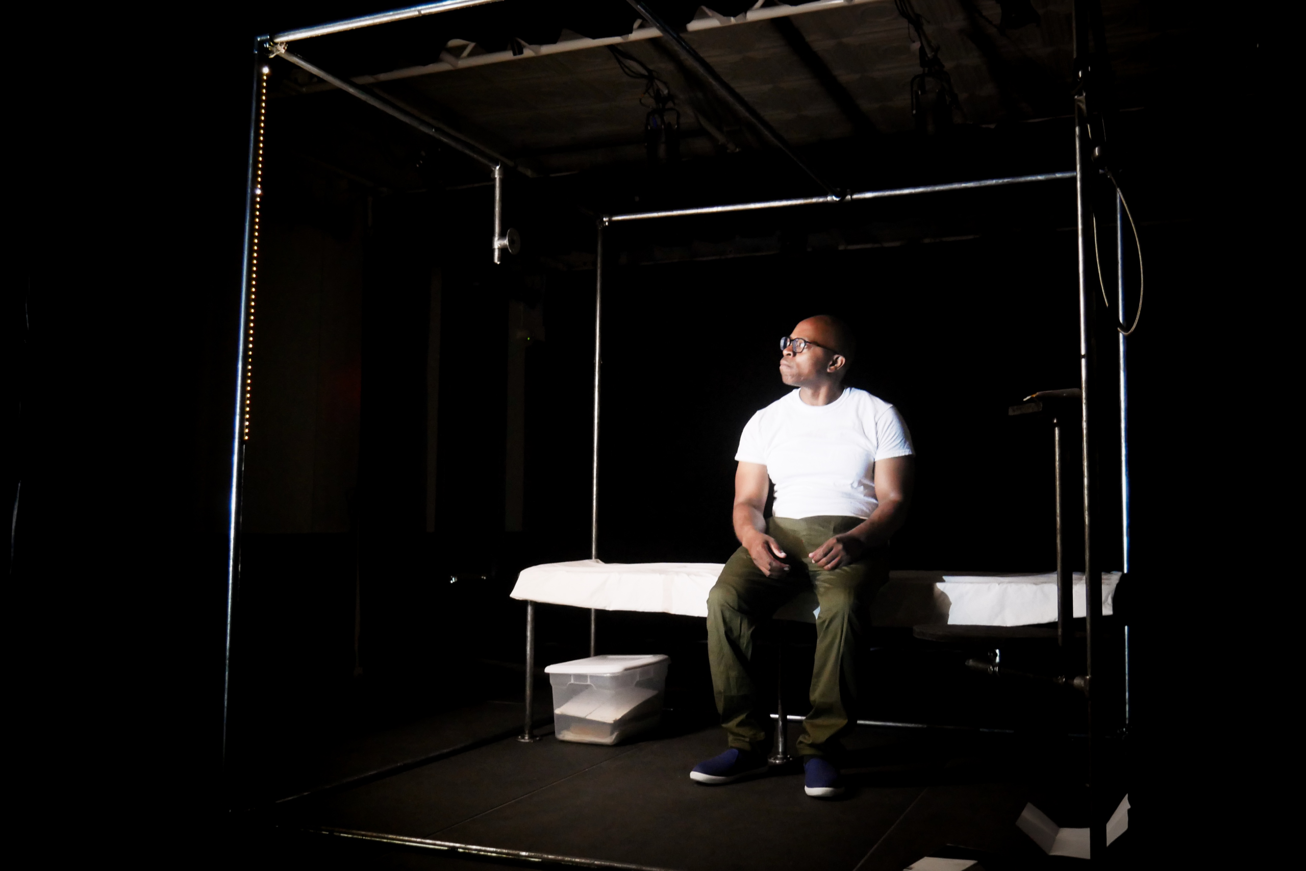 A man in a white shirt sits on a white mattress looking to the distance.